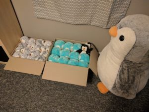 A very large stuffed penguin, Hoogerheide, looks out over two boxes of smaller stuffed penguins. The first box is primarily a light teal, with one smaller black-and-white penguin in the corner, and the one on the right is full of smaller gray penguins.