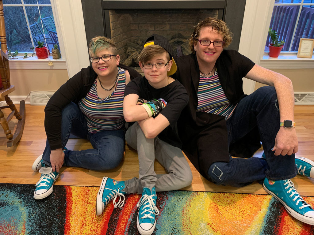 Three people in brightly colored clothes are seen sitting on the floor in front of a fireplace, laughing.