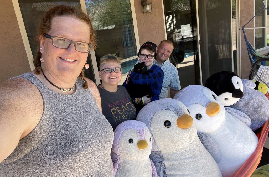 Four people are standing behind a hammock with four large stuffed penguins and a stuffed fish in it. One person is holding a cat.